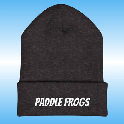 A dark charcoal gray heather knit beanie with a folded cuff featuring the words 'PADDLE FROGS' embroidered in white. The beanie is centered against a light blue gradient background.