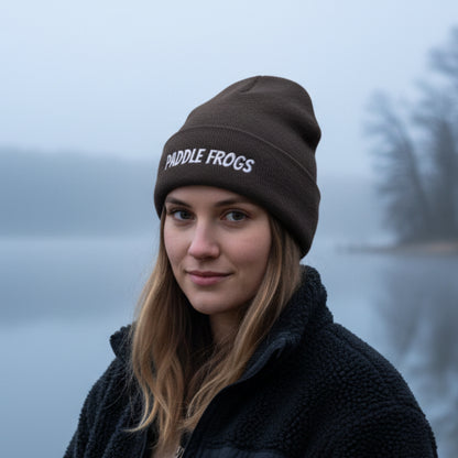 A woman wearing a dark grey beanie with 'PADDLE FROGS' embroidered in white on the front, photographed outdoors against a misty lake background.