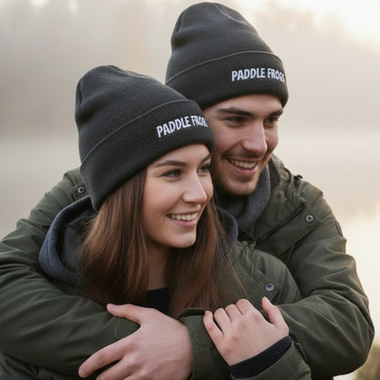 A smiling couple outdoors wearing matching dark gray knit beanie hats with white 'PADDLE FROGS' text on the front cuff. The woman is in front and the man is behind her, both wearing olive green jackets.