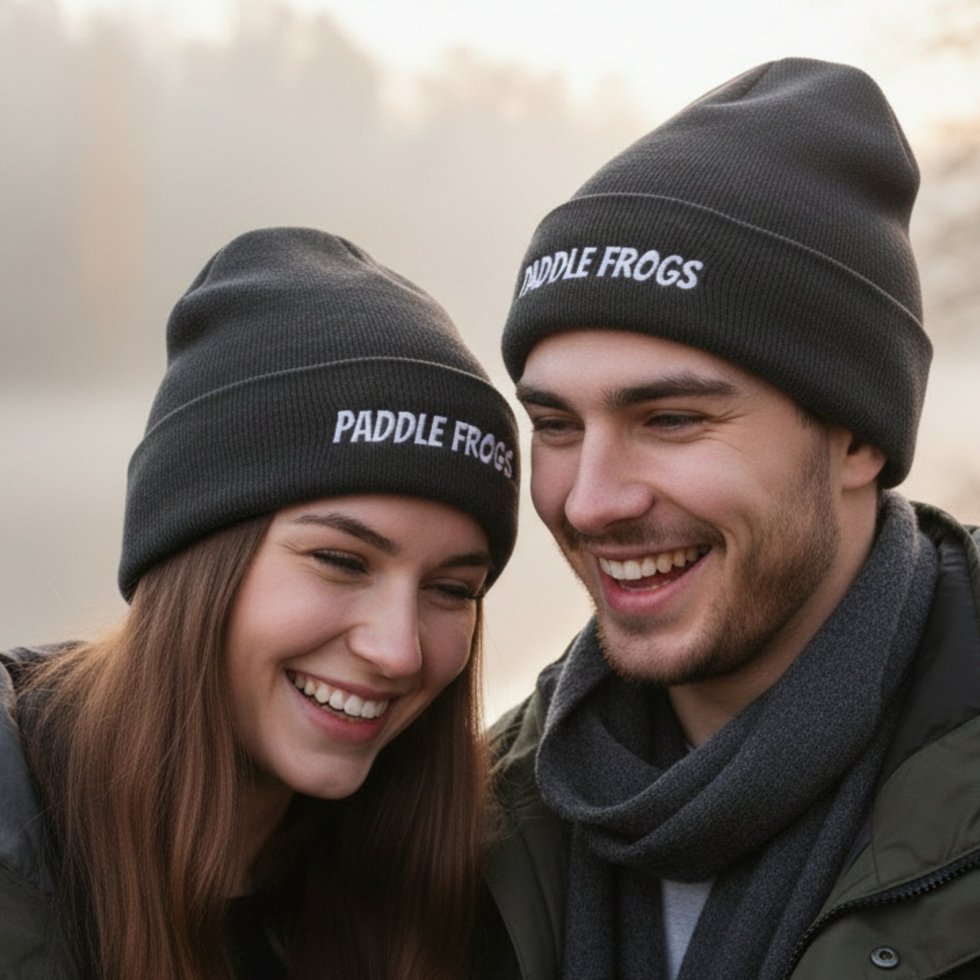 Two smiling people outdoors wearing dark knit beanie hats with white embroidered text reading 'PADDLE FROGS'.