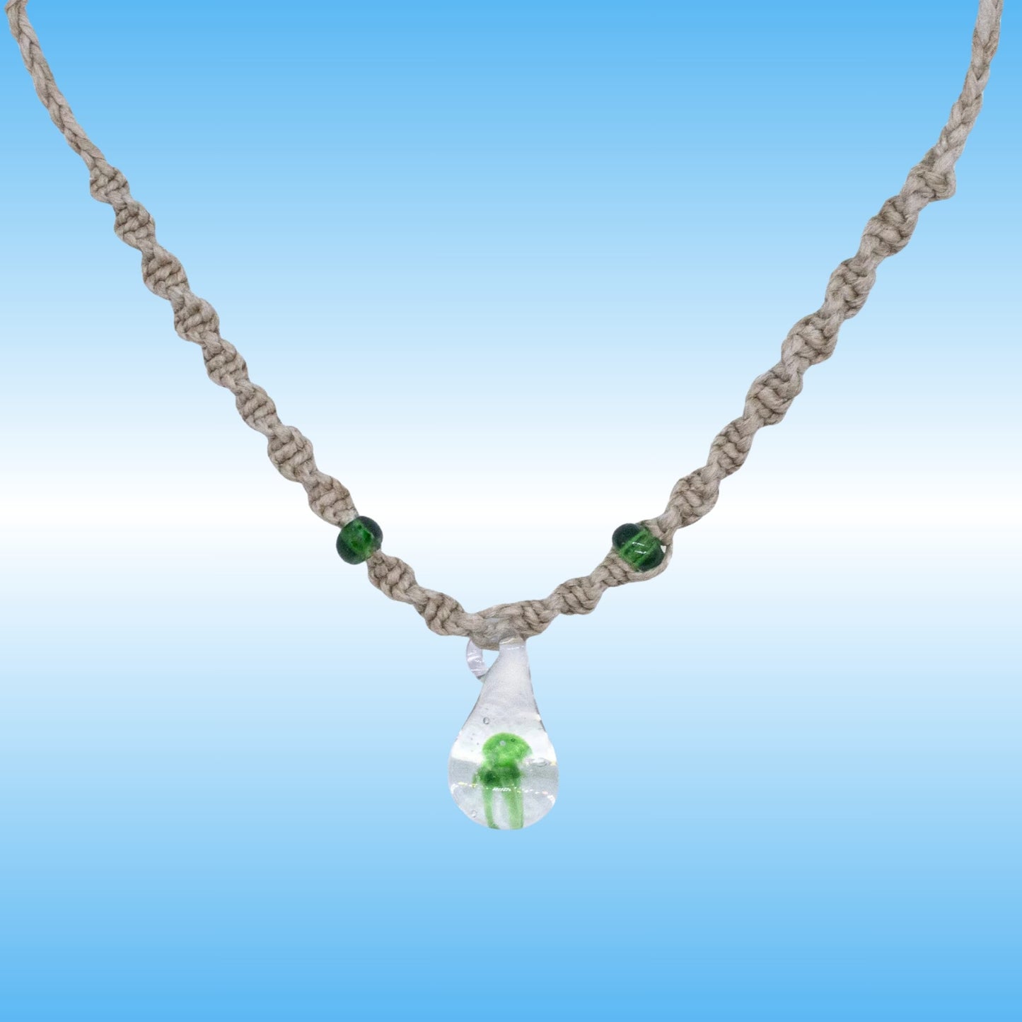 A handmade macrame hemp necklace with a clear glass teardrop pendant containing a small green mushroom, accented with two green beads on a blue gradient background.