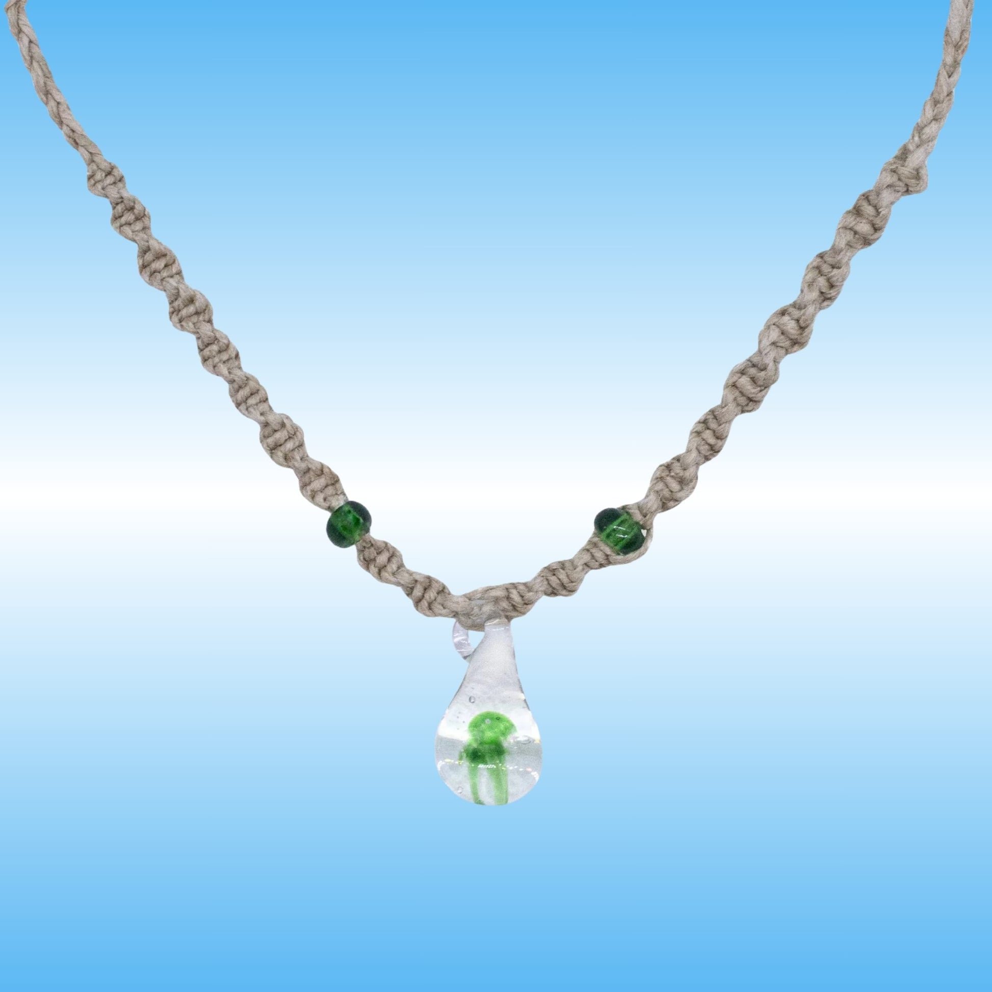 A handmade macrame hemp necklace with a clear glass teardrop pendant containing a small green mushroom, accented with two green beads on a blue gradient background.