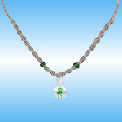 A handmade macrame hemp necklace with a clear glass teardrop pendant containing a small green mushroom, accented with two green beads on a blue gradient background.