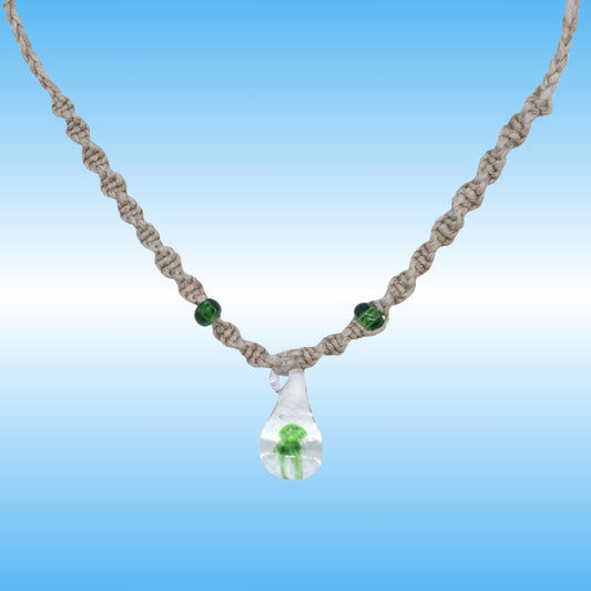A handmade macrame hemp necklace with a clear glass teardrop pendant containing a small green mushroom, accented with two green beads on a blue gradient background.