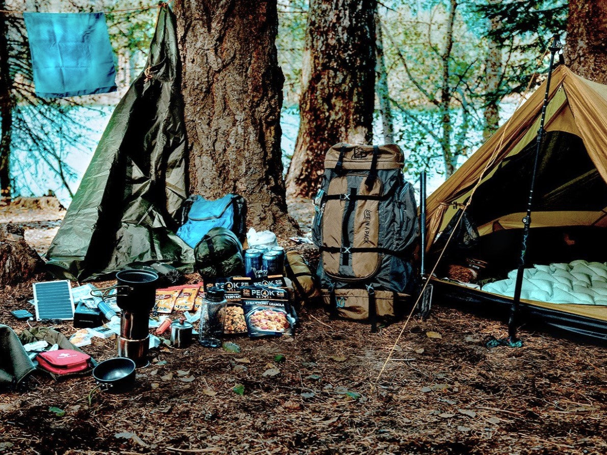 A forest camping setup with a tan tent on the right, a dark green tarp on the left, a large hiking backpack in the center, and camping gear laid out on the ground including food, a stove, bowls, and a small solar panel.