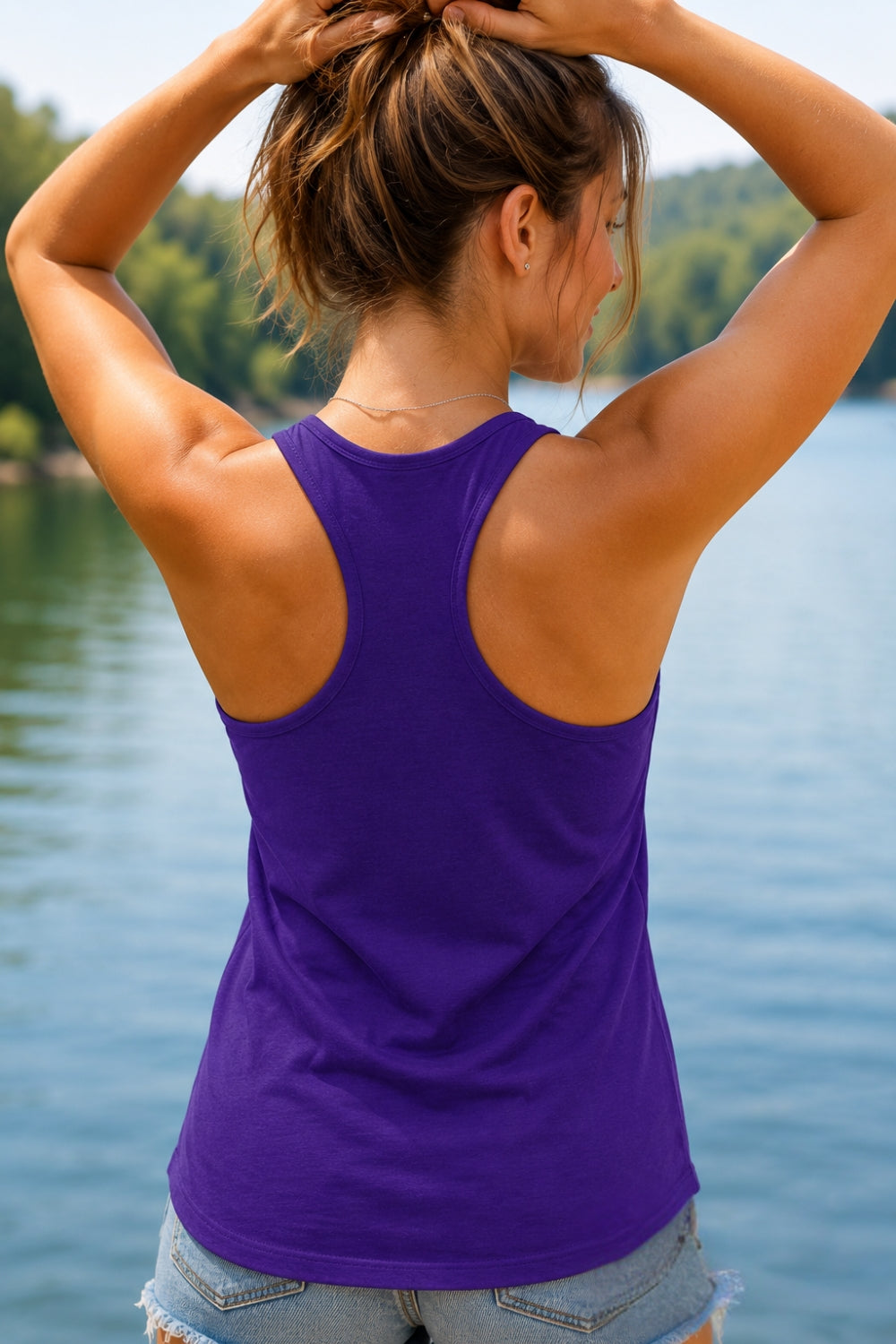 A woman viewed from behind wearing a purple rush Paddle Frogs racerback tank top, standing outdoors by a lake with trees in the background.