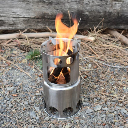 A small stainless steel bio fuel camp stove with visible flames burning inside, placed outdoors on gravel with pine needles and a log in the background.