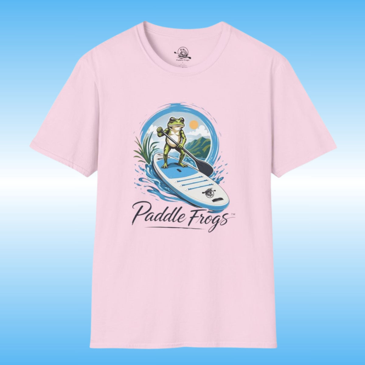 A light pink short-sleeve t-shirt with a graphic of a frog paddleboarding on the front, with the text 'Paddle Frogs' printed below the illustration. The t-shirt is shown against a blue gradient background.