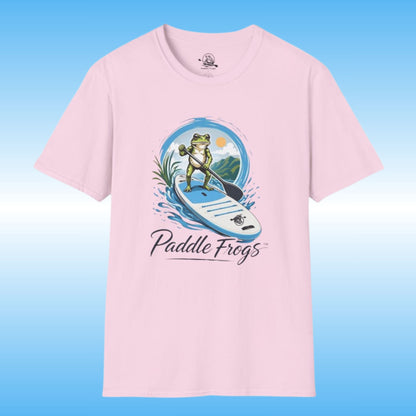 A light pink short-sleeve t-shirt with a graphic of a frog paddleboarding on the front, with the text 'Paddle Frogs' printed below the illustration. The t-shirt is shown against a blue gradient background.
