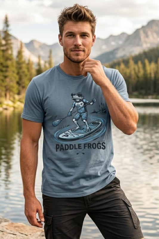 A man wearing a stone blue graphic t-shirt with a frog paddleboarding design and the text 'PADDLE FROGS', standing by a lake with mountains in the background.