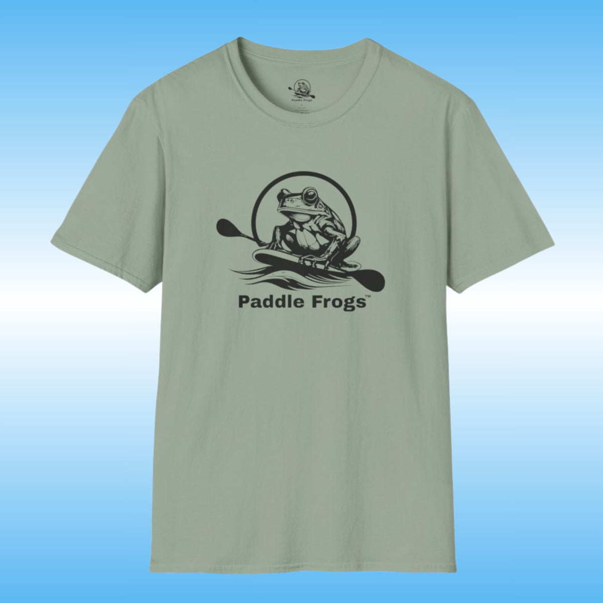A sage green crewneck t-shirt featuring a black graphic of a frog on a paddle board with the text 'Paddle Frogs' printed on the chest, set against a blue gradient background.