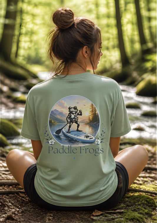 A woman sitting on a mossy log in a forest, viewed from behind, wearing a sage green t-shirt with a large graphic of a frog on a paddleboard and the text 'Paddle Frogs' printed on the back.