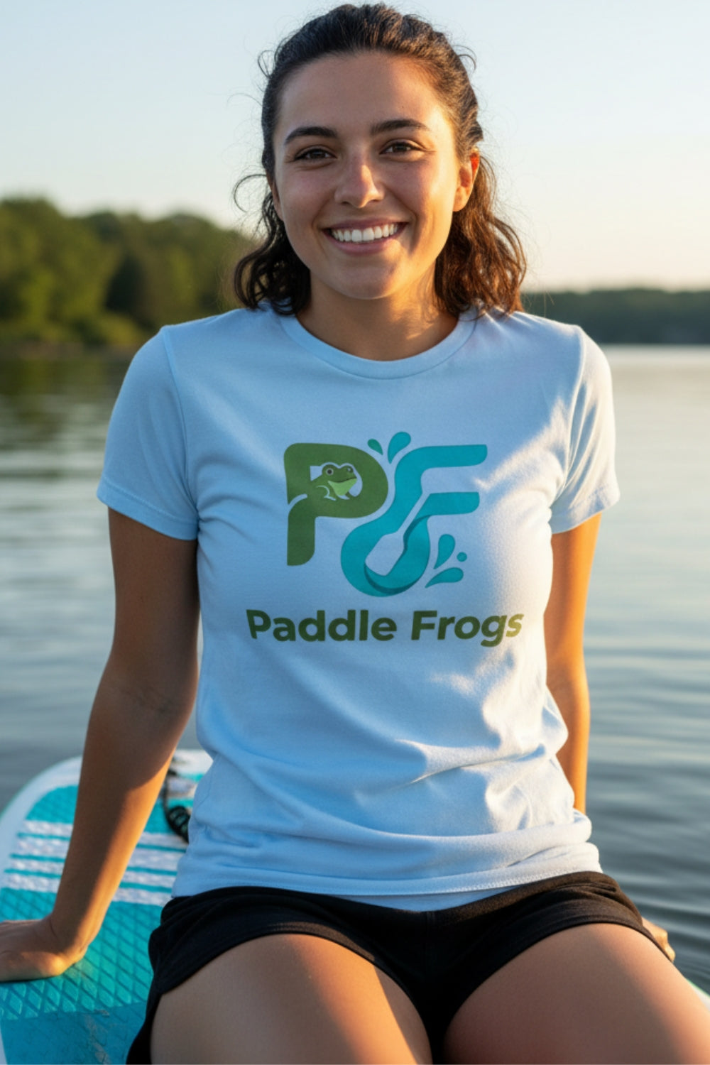 A smiling woman wearing a light blue t-shirt with a green and teal 'Paddle Frogs' logo, sitting on a paddleboard on a lake with trees in the background.