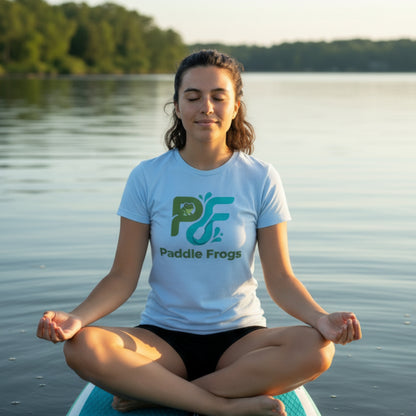 A woman meditating cross-legged on a paddleboard on a calm lake, wearing a light blue t-shirt with a green and black 'Paddle Frogs' logo.