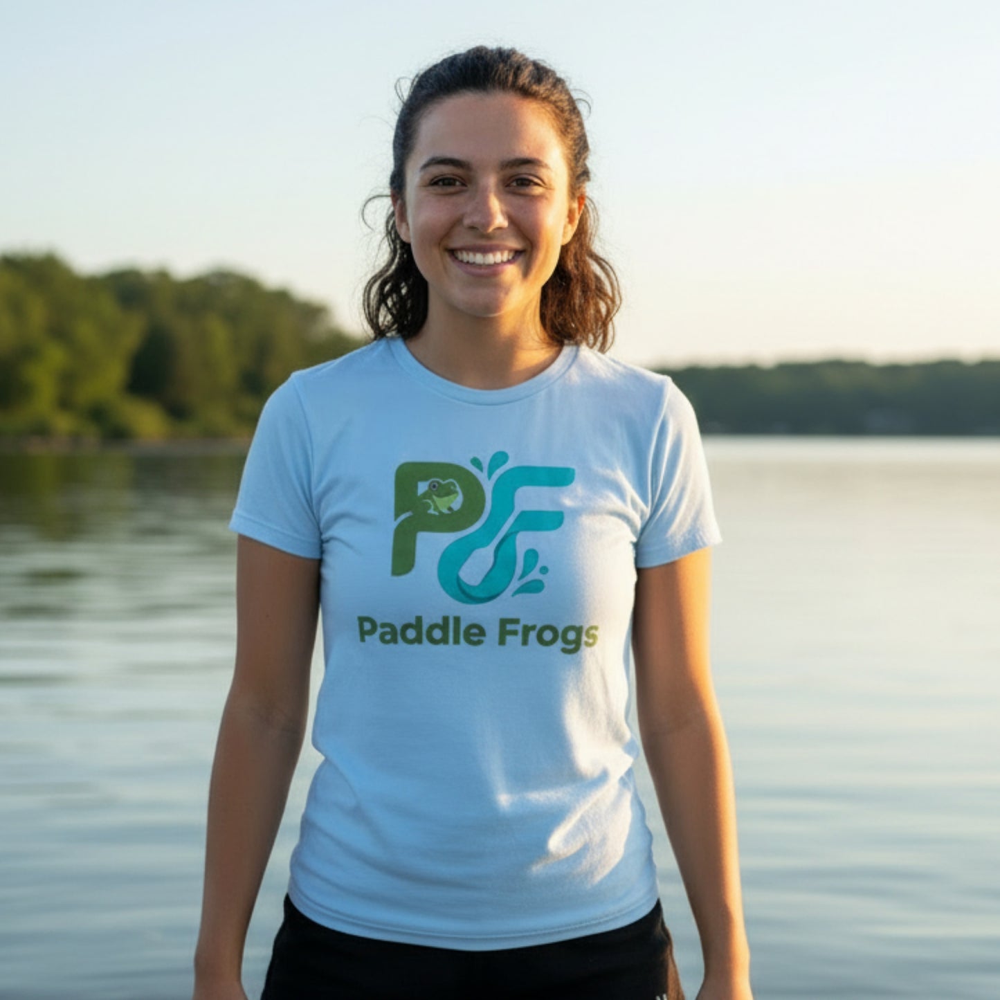 A smiling woman stands on her paddleboard at a calm lake wearing a light blue T-shirt with a large green and teal 'Paddle Frogs' logo and text on the front.
