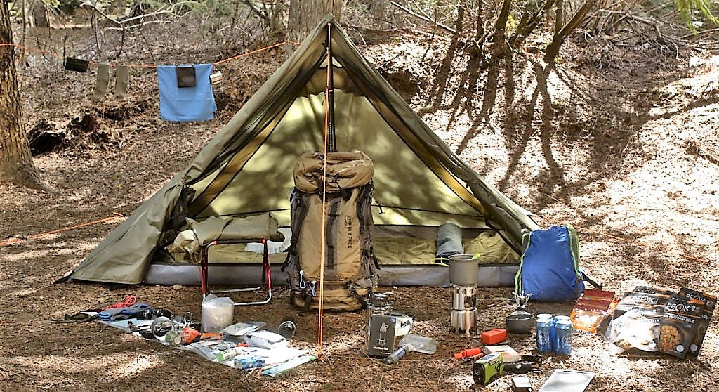 An olive green A-frame camping tent pitched in a wooded outdoor setting, surrounded by camping gear including a large backpack, blue sleeping bag, folding chair, stove, cups, and other camping supplies on the ground.