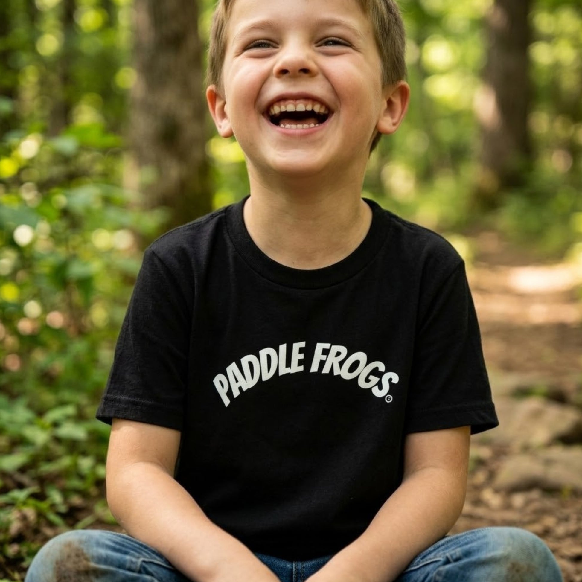 A young boy laughing outdoors in a black t-shirt with 'PADDLE FROGS' printed in white arched text across the chest, set against a blurred forest background.