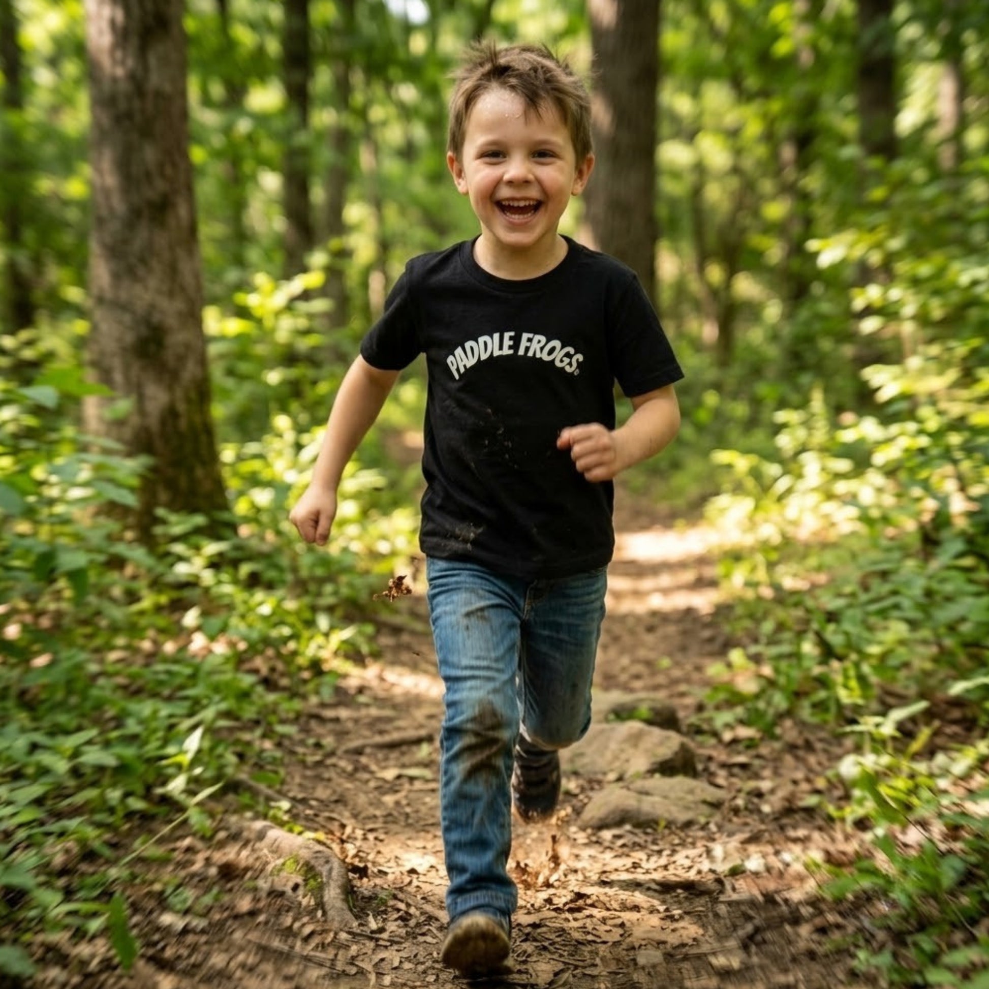 A young boy smiling and running on a forest path wearing a black t-shirt with 'PADDLE FROGS' printed in white across the chest, paired with blue jeans.