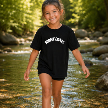 A young girl smiling while walking through a shallow outdoor stream, wearing a black short-sleeve T-shirt with 'PADDLE FROGS' printed in white across the chest.