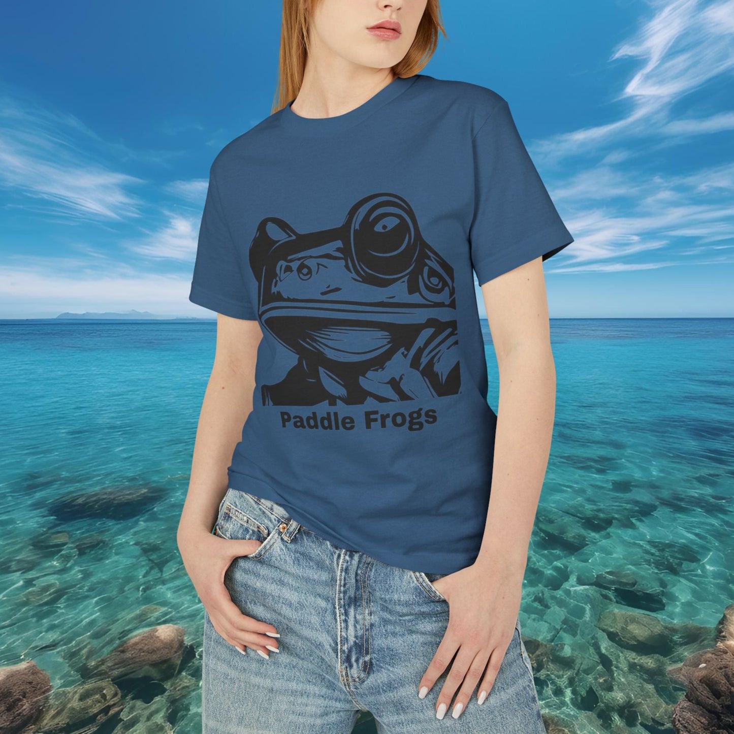 Person wearing a Faded Navy blue t-shirt with a frog graphic and 'Paddle Frogs' text, standing in front of a scenic ocean view.
