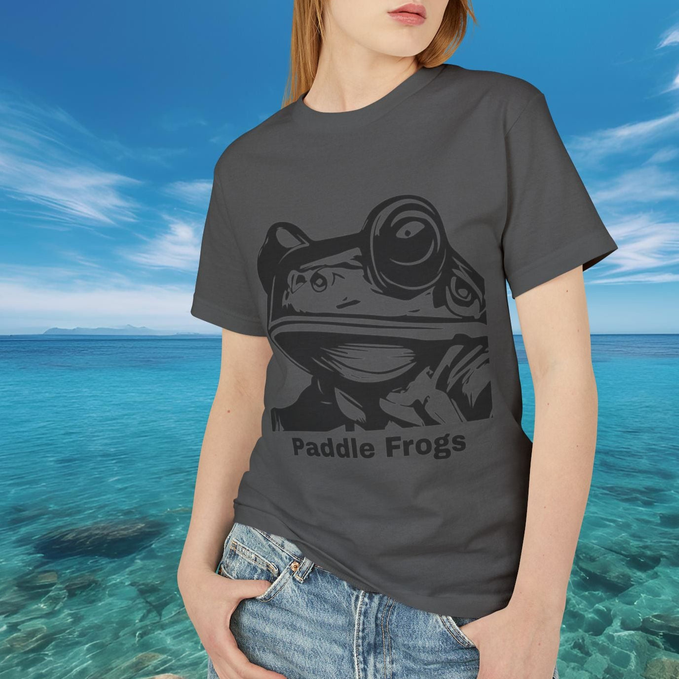 Person wearing a faded black t-shirt with a frog graphic and 'Paddle Frogs' text, standing in front of a scenic ocean view.