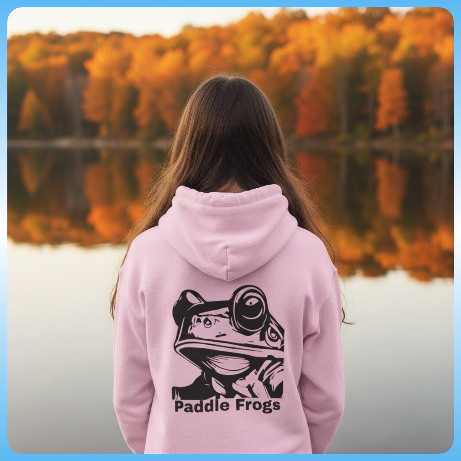 Person wearing a pink hoodie with 'Paddle Frogs' design, standing by a lake with autumn trees.