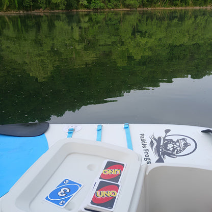 A close-up view from a paddle board on calm water, showing the front deck area with a white storage tray, blue straps, a black paddle logo reading Paddle Frogs, and two UNO playing cards. Trees and reflective water fill the background.