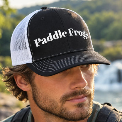 A man wearing a black and white trucker hat with 'Paddle Frogs' embroidered in white on the front panel, photographed outdoors with a blurred natural background.