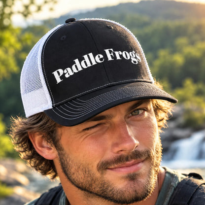 A man outdoors wearing a black and white trucker hat with 'Paddle Frogs' embroidered in white on the front panel, with a blurred forest and waterfall background.