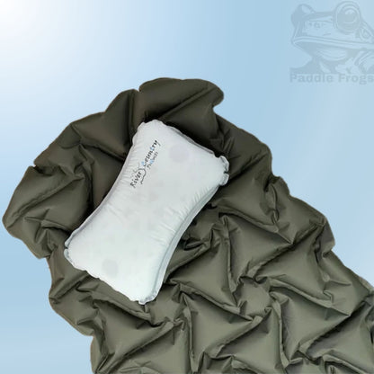 A grey inflatable sleeping pillow with the 'Paddle Frog' logo, shown resting on top of an olive green inflatable soft packable sleeping pad against a light blue gradient background.