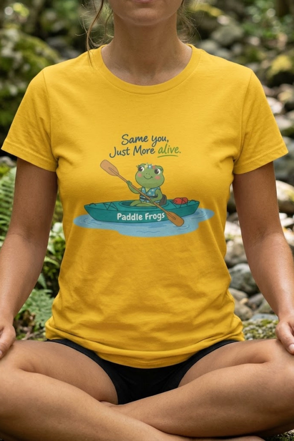 A woman wearing a bright yellow graphic t-shirt sits crisscross applesauce outdoors in a natural setting. The t-shirt features a cartoon frog paddling in a teal kayak with the text 'Same you, Just More alive above the frog and Paddle Frogs on the kayak.