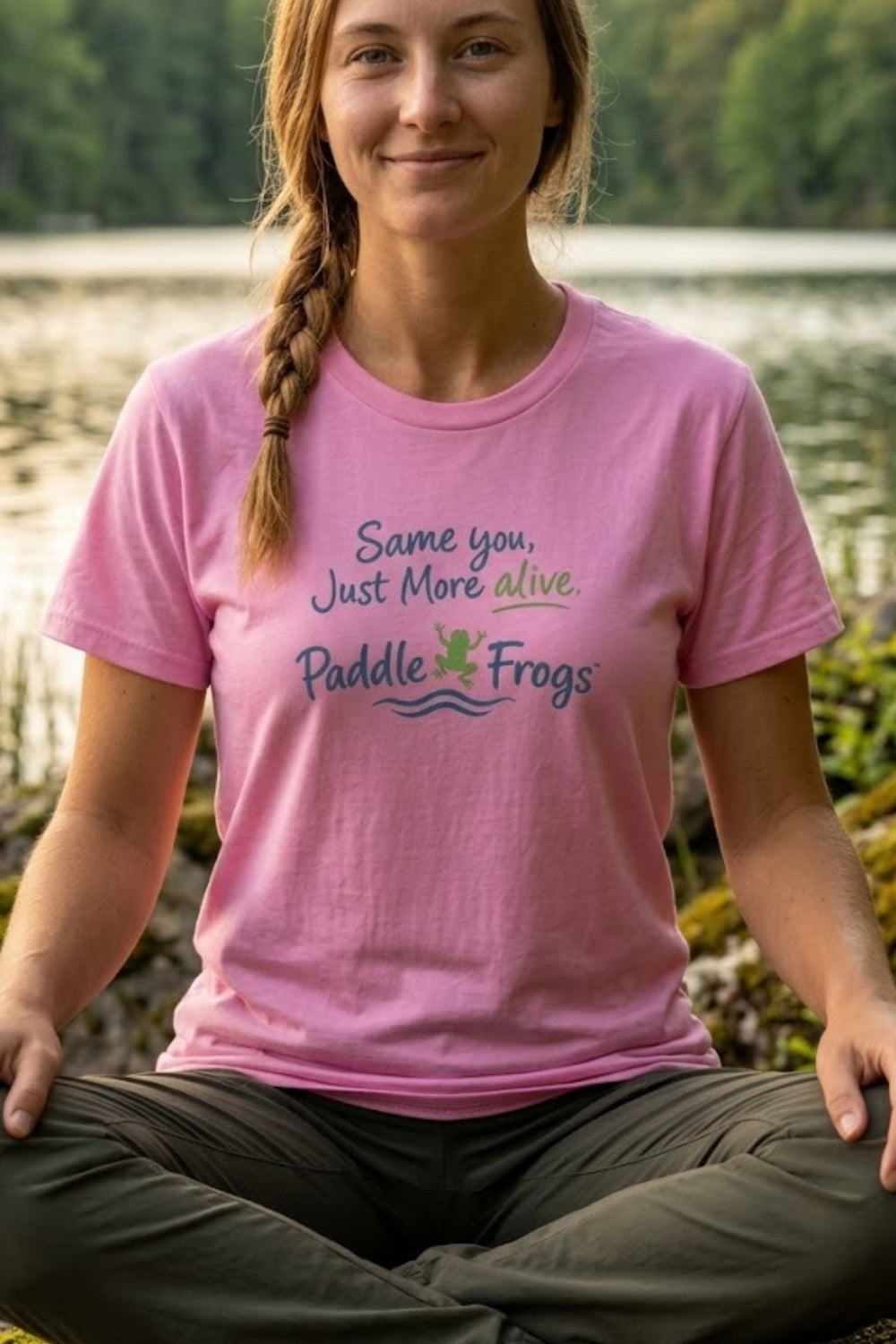 A woman sitting cross-legged outdoors by a lake wears a pink graphic T-shirt that reads 'Same you, Just More alive. Paddle Frogs' with a small frog logo.
