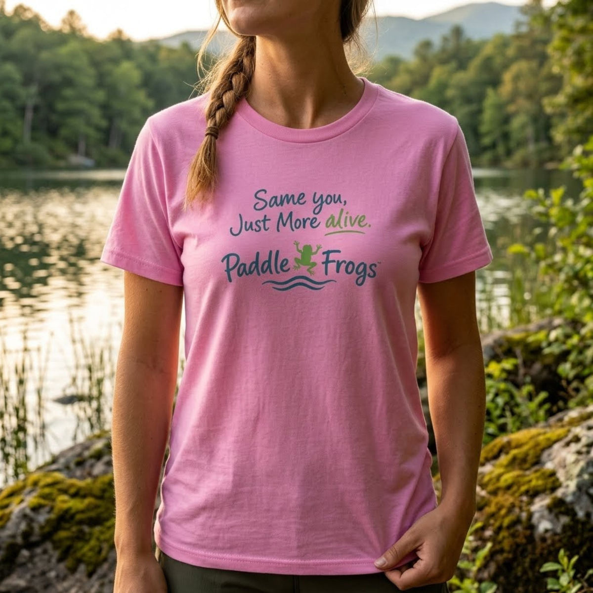 A woman wearing a pink short-sleeve t-shirt with a graphic that reads 'Same you, Just More alive. Paddle Frogs' in green and blue lettering, standing outdoors in front of a scenic lake and forest background.