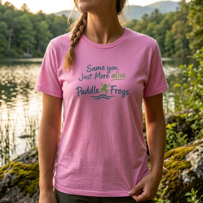 A woman wearing a pink short-sleeve t-shirt with a graphic that reads 'Same you, Just More alive. Paddle Frogs' in green and blue lettering, standing outdoors in front of a scenic lake and forest background.
