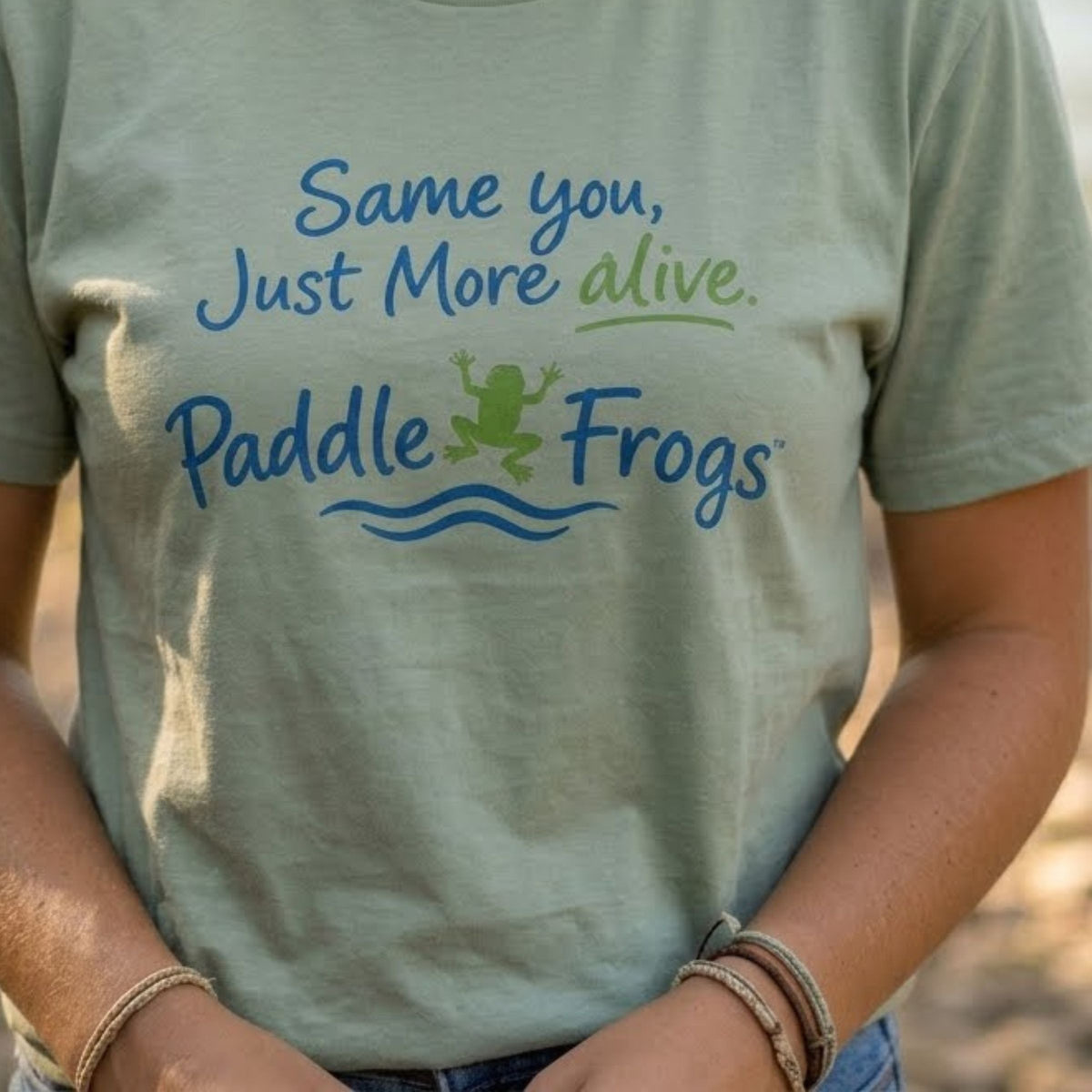 A person wearing a sage green graphic t-shirt with the text 'Same you, Just More alive.' and 'Paddle Frogs' in blue and green, with a frog jumping over blue waves.