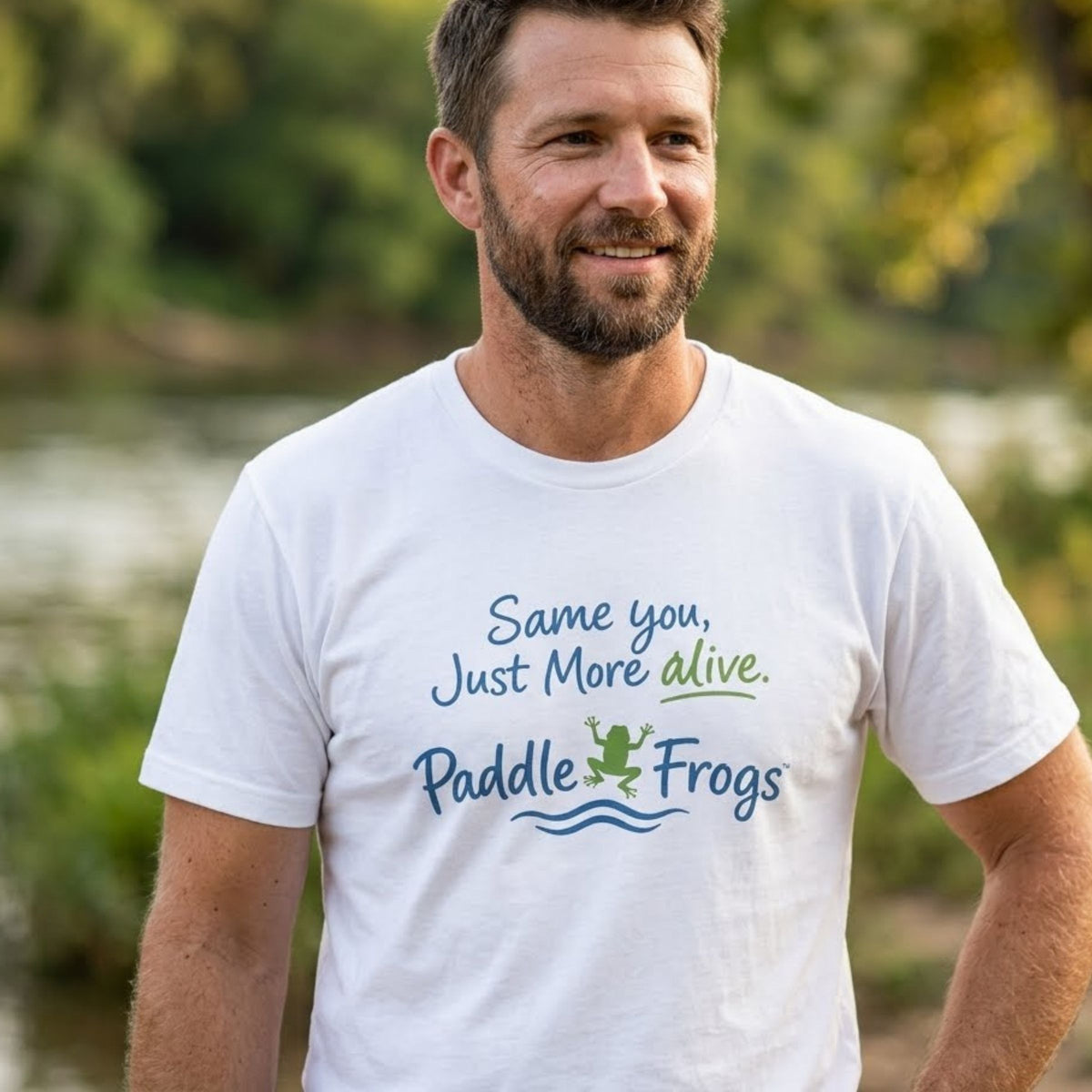 A man wearing a white graphic t-shirt with the text 'Same you, Just More alive. Paddle Frogs' and a small frog illustration, photographed outdoors in front of a blurred green background.
