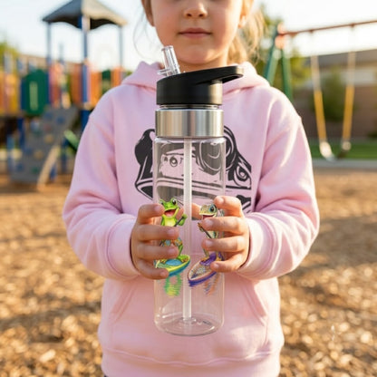 A child in a pink hoodie holds a clear plastic water bottle with a black lid and straw in an outdoor playground setting. The bottle has a silver collar and colorful cartoon frog and lizard paddle board graphics.