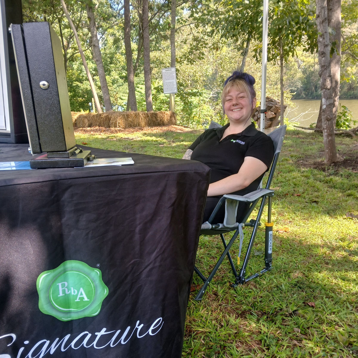 A woman smiling while sitting in a folding camp chair next to a black table draped with a black cloth featuring a green 'Rba Signature' logo, set outdoors in a grassy shaded area in a park like setting with trees and the Emory River in the background. Located in Harriman, TN at Paddle Frogs Sunday Market.