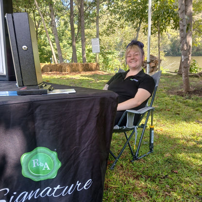 A woman smiling while sitting in a folding camp chair next to a black table draped with a black cloth featuring a green 'Rba Signature' logo, set outdoors in a grassy shaded area in a park like setting with trees and the Emory River in the background. Located in Harriman, TN at Paddle Frogs Sunday Market.