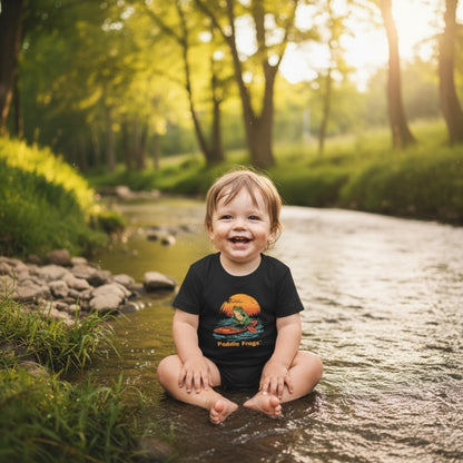 Sitting crisscross applesauce in the creek wearing the black Sunset Frog Paddle Board Explorer Toddler Tee "Skate & Paddle Frogs™ 