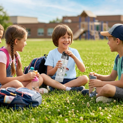 Three children sitting on green grass outdoors near a school building, each holding a reusable water bottle. A fourth child is partially visible on the left, and backpacks are on the ground nearby.