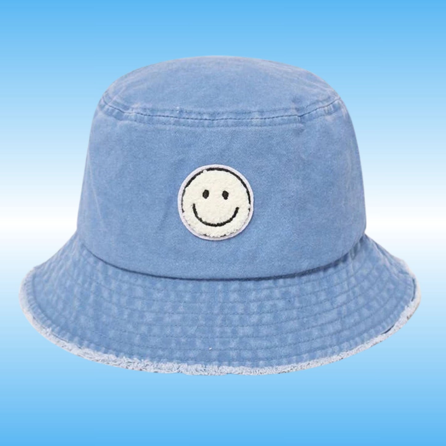 A light blue denim bucket hat with a white embroidered smiley face patch on the front, set against a light blue gradient background.