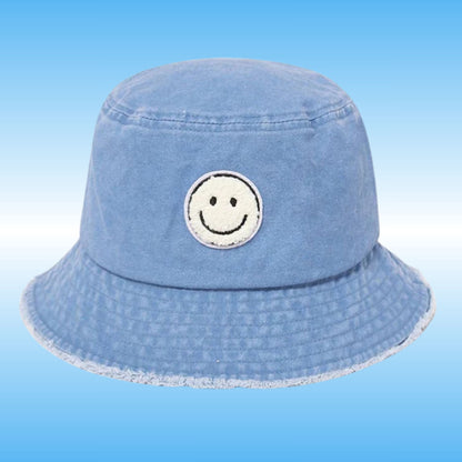 A light blue denim bucket hat with a white embroidered smiley face patch on the front, set against a light blue gradient background.