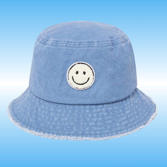 A light blue denim bucket hat with a white embroidered smiley face patch on the front, set against a light blue gradient background.