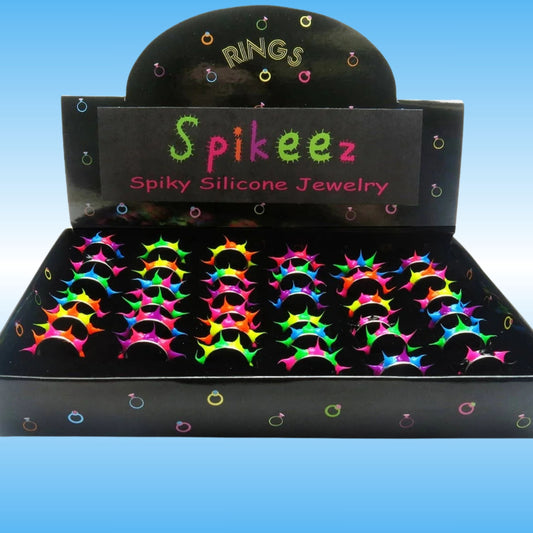 A black display box labeled 'Spikeez Spiky Silicone Jewelry' filled with multiple pairs of neon rainbow-colored spiked rings, shown against a blue gradient background.