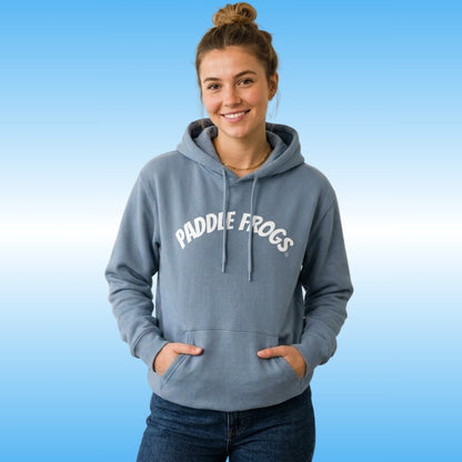 A woman wearing a stone blue hoodie with a retro white 'PADDLE FROGS' printed in white arched text across the chest, posed against a light blue gradient background.