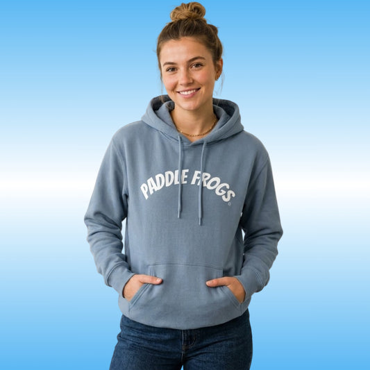A woman wearing a stone blue hoodie with a retro white 'PADDLE FROGS' printed in white arched text across the chest, posed against a light blue gradient background.