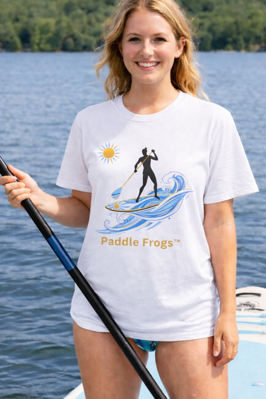 A smiling blonde woman standing the Divine Escape stand-up paddleboard on a lake, wearing a white t-shirt with a sun and paddleboarder graphic that says "Paddle Frogs." She is holding a black paddle.