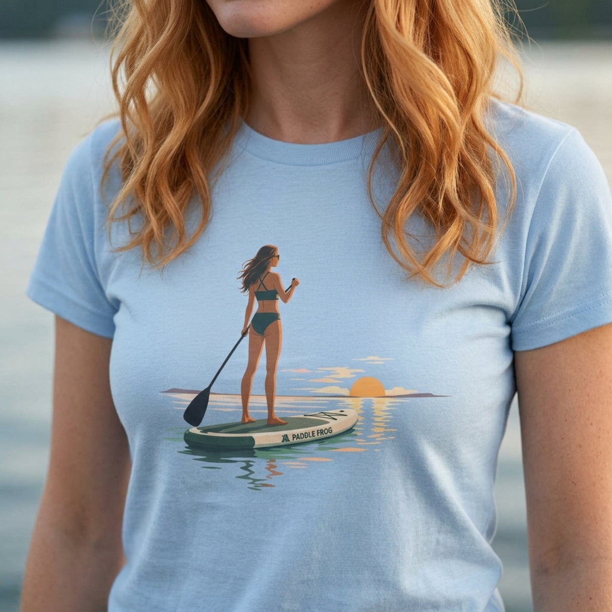A woman wearing a light blue ladies SUP crewneck t-shirt with a graphic print of a woman paddleboarding on water at sunset. The background is a soft-focus outdoor water setting. Paddle Frogs Outdoor Gear 