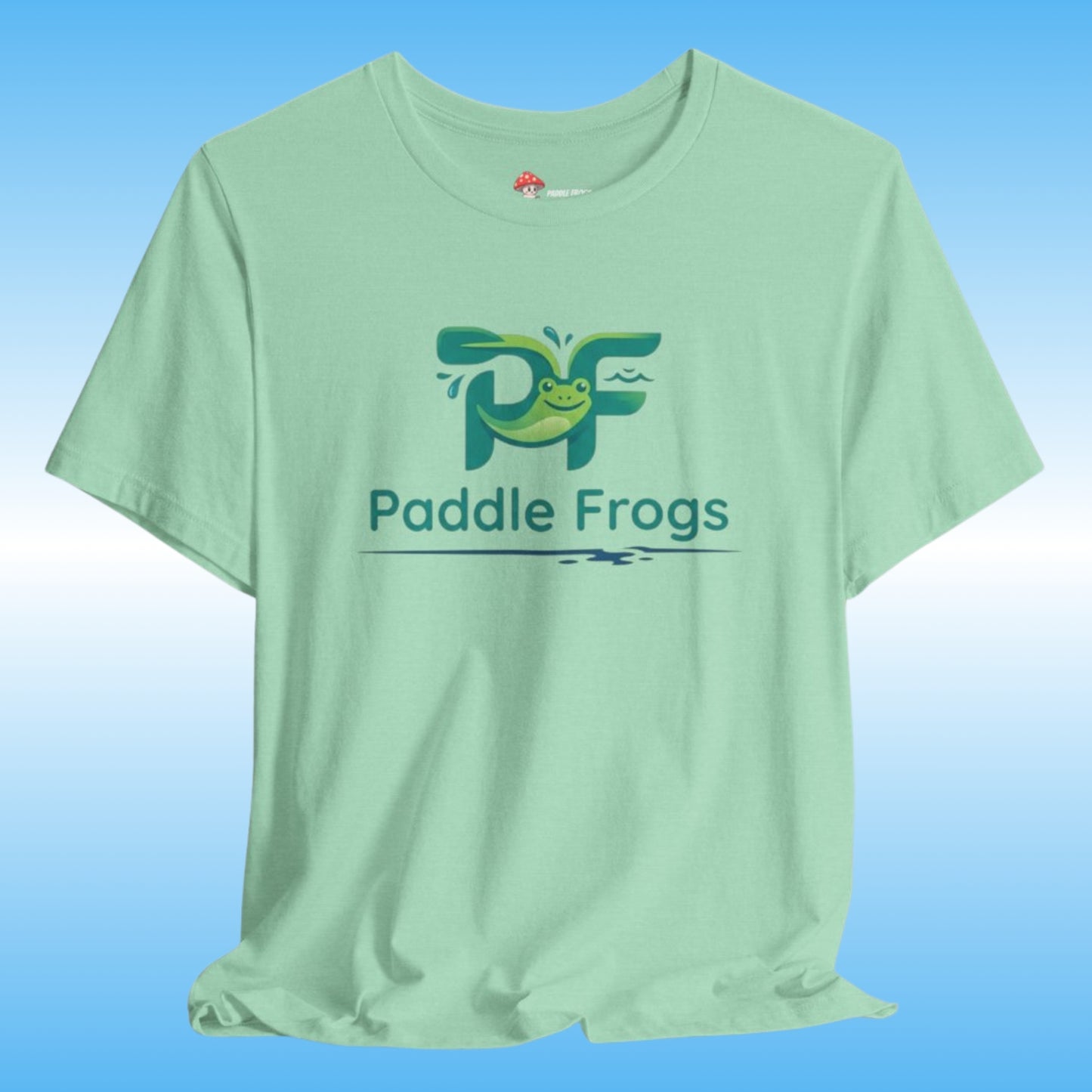 A light mint green t-shirt featuring a green and blue 'Paddle Frogs' tadpole printed on the chest, displayed against a blue gradient background.